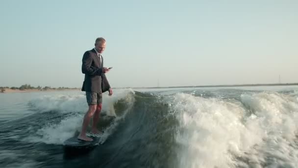 Ένας σέρφερ που πηδάει σε μια σανίδα με ένα κοστούμι με ένα τηλέφωνο στα χέρια του. Ένας έμπειρος wakeboarder ψεκάζει νερό σταγόνες στην κάμερα. — Αρχείο Βίντεο