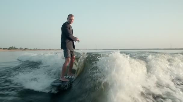 Um surfista a saltar num wakeboard de fato com um telefone nas mãos. Um wakeboarder experiente pulveriza água cai na câmera. — Vídeo de Stock