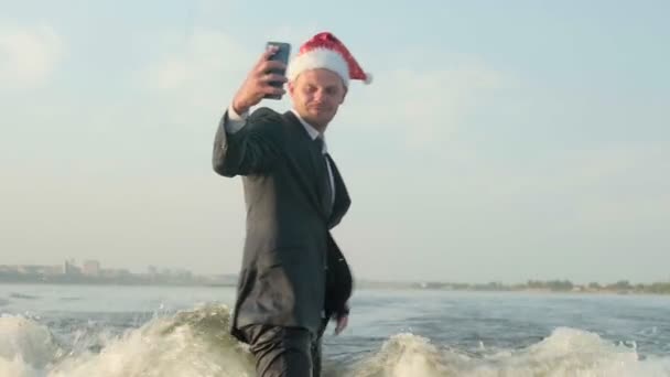 Surfař skákající na wakeboardu v obleku a s vánočním červeným kloboukem na hlavě je vyfocen na telefonu. Zkušený wakeboarder rozstřikuje do kamery kapky vody. — Stock video