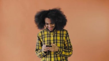 Turuncu arka planda Afrikalı saç stili olan genç bir adam telefona bakar ve biriyle mutlu mesut mesajlaşır. Renkli bir arkaplanda duygular