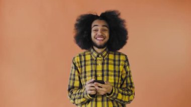 Turuncu arka planda Afrikalı saç stili olan genç bir adam telefona bakar ve mutlu olur. Renkli bir arkaplanda duygular