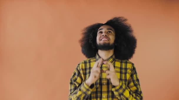 Młody mężczyzna z afrykańską fryzurą na pomarańczowym tle krzyżuje palce i modli się. Emocje na kolorowym tle. — Wideo stockowe