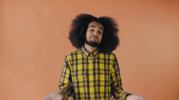 Młody mężczyzna z afrykańską fryzurą na pomarańczowym tle wzrusza ramionami. Emocje na kolorowym tle. — Wideo stockowe