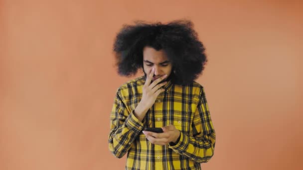 Un joven con un peinado africano sobre un fondo naranja mira el teléfono y está felizmente sorprendido. Emociones sobre un fondo coloreado — Vídeos de Stock