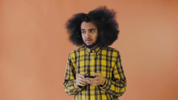 Un joven con un peinado africano sobre un fondo naranja mira el teléfono y las dudas. Emociones sobre un fondo coloreado — Vídeos de Stock