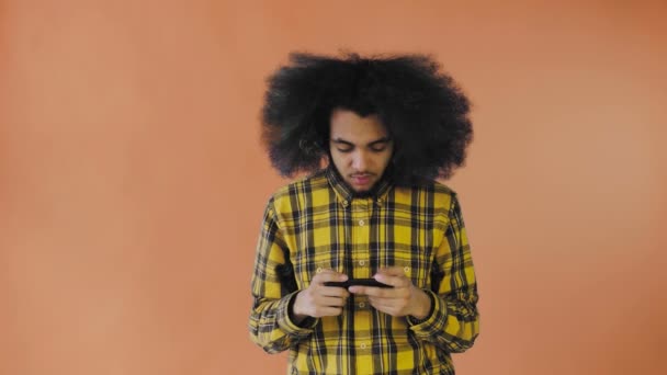 Un joven con un peinado africano sobre un fondo naranja está jugando por teléfono. Sobre un fondo de color — Vídeo de stock