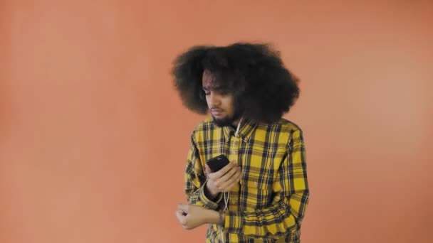 Mladý muž s africkým účesem na oranžovém pozadí poslouchá hudbu a sluchátka. Na barevném pozadí — Stock video