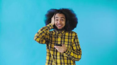 Mavi arka planda Afrikalı saç stili olan genç bir adam telefona bakar ve mutlu bir şekilde şaşırır. Renkli bir arkaplanda duygular