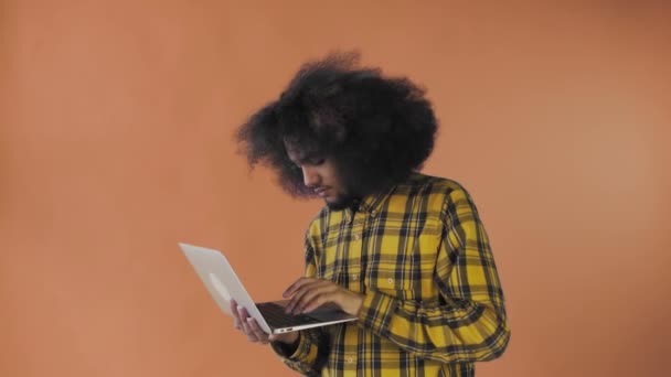 Um jovem com um penteado africano em um fundo laranja está digitando em um laptop. Em um fundo colorido — Vídeo de Stock