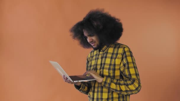 Ένας νεαρός με αφρικάνικο χτένισμα σε πορτοκαλί φόντο πληκτρολογεί σε ένα λάπτοπ. Σε έγχρωμο φόντο — Αρχείο Βίντεο