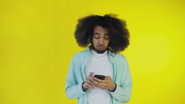 Egy afrikai frizurájú, narancssárga hátterű fiatalember ránéz a telefonra és boldog. Érzelmek egy színes háttér — Stock videók