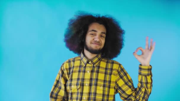 Un jeune homme avec une coiffure africaine sur fond bleu montre le geste OK. Émotions sur un fond coloré. — Video
