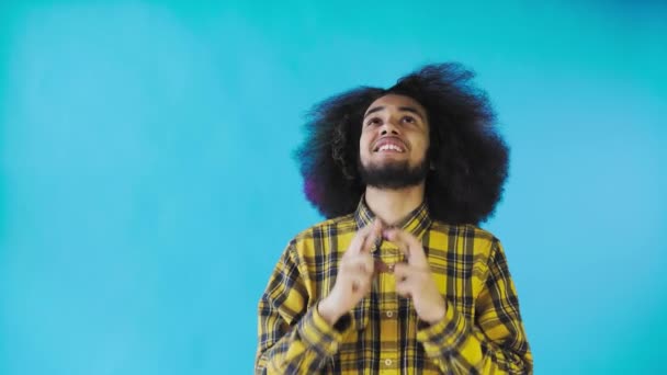 Ein junger Mann mit afrikanischer Frisur auf blauem Grund drückt ihm die Daumen und betet. Emotionen auf farbigem Hintergrund. — Stockvideo