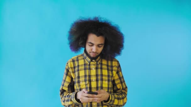 Un joven con un peinado africano sobre un fondo azul está hablando en su teléfono. — Vídeos de Stock