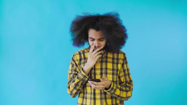 Um jovem com um penteado africano em um fundo azul olha para o telefone e fica feliz surpreso. Emoções em um fundo colorido — Vídeo de Stock