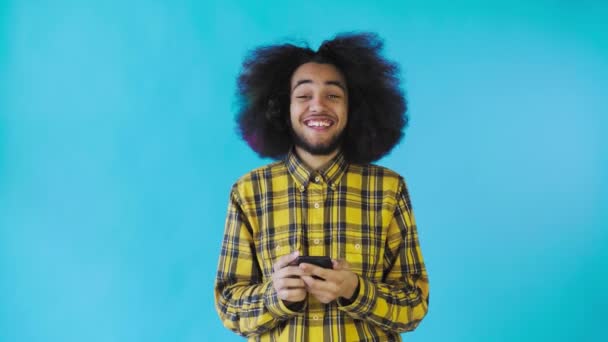 Un jeune homme avec une coiffure africaine sur fond bleu regarde le téléphone et est heureux. Émotions sur un fond coloré — Video