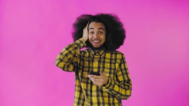 Pembe arka planda Afrikalı saç stili olan genç bir adam telefona bakar ve mutlu bir şekilde şaşırır. Renkli bir arkaplanda duygular