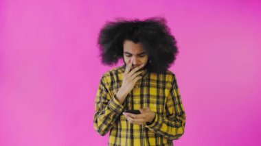 Pembe arka planda Afrikalı saç stili olan genç bir adam telefona bakar ve mutlu bir şekilde şaşırır. Renkli bir arkaplanda duygular
