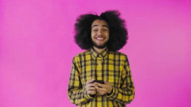 Pembe arka planda Afrikalı saç stili olan genç bir adam telefona bakar ve mutludur. Renkli bir arkaplanda duygular