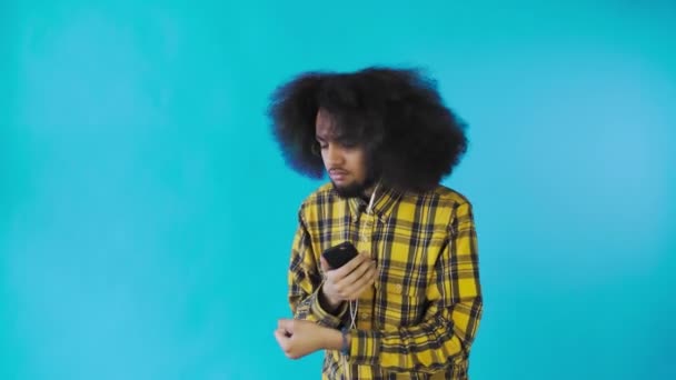 Seorang pemuda dengan gaya rambut Afrika dengan latar belakang biru mendengarkan musik dan headphone. Pada latar belakang berwarna — Stok Video