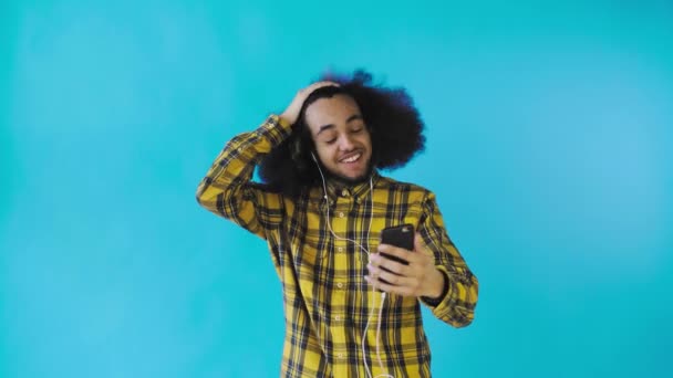 Un joven con un peinado africano sobre un fondo azul en auriculares se comunica a través de un enlace de vídeo. Sobre un fondo de color — Vídeos de Stock