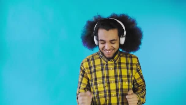 En ung man med en afrikansk frisyr på blå bakgrund lyssnar på musik och hörlurar. På en färgad bakgrund — Stockvideo