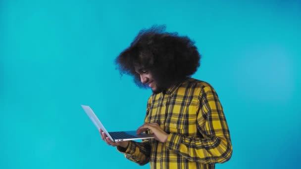 Um jovem com um penteado africano em um fundo azul está digitando em um laptop. Em um fundo colorido — Vídeo de Stock