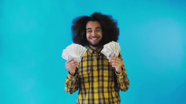Um homem afro-americano feliz segurando um fã de notas em suas mãos e olhando para a câmera, de pé isolado em um fundo azul — Vídeo de Stock