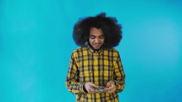 Un hombre afroamericano feliz sosteniendo billetes en sus manos, contándolos y mirando a la cámara, parado aislado sobre un fondo azul — Vídeos de Stock