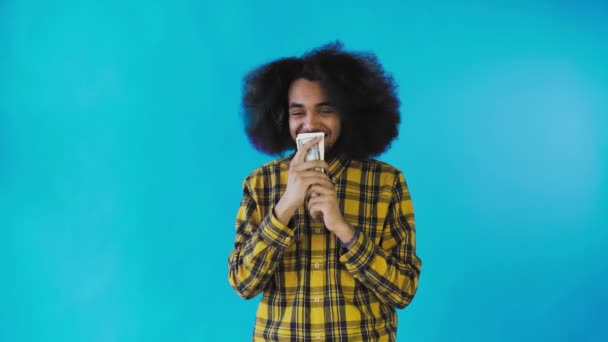 Um homem afro-americano feliz segurando notas em suas mãos, beijando-as e olhando para a câmera, fica isolado em um fundo azul — Vídeo de Stock