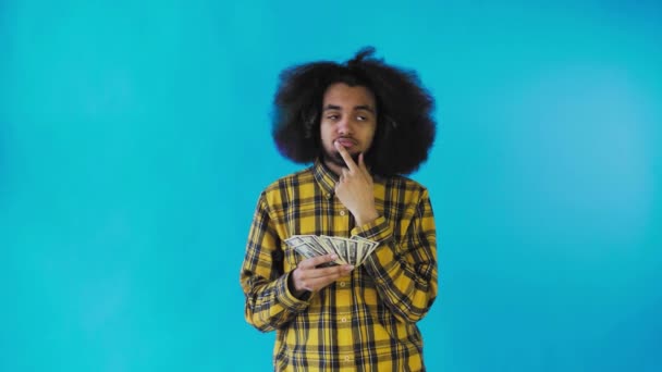 Um afro-americano feliz segurando dinheiro em suas mãos e pensando sobre o que gastá-lo, fica isolado em um fundo azul — Vídeo de Stock