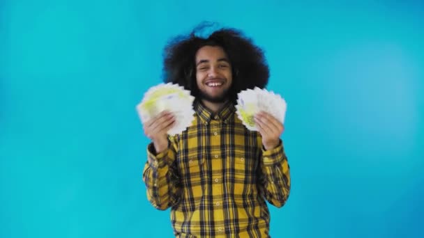 Een gelukkige Afro-Amerikaanse man die een fan van bankbiljetten in zijn handen houdt en naar de camera kijkt, geïsoleerd op een blauwe achtergrond — Stockvideo