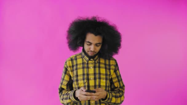 一个有着粉色背景的非洲发型的年轻人正在对着他的手机讲话. — 图库视频影像