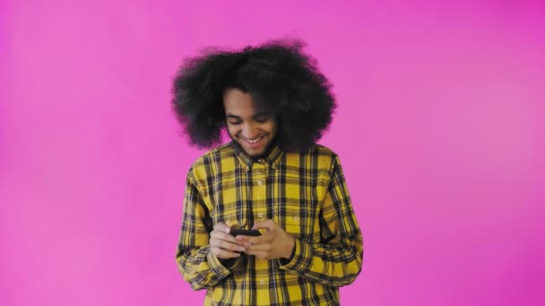 En ung man med en afrikansk frisyr på rosa bakgrund tittar på telefonen och messar gladeligen med någon. Känslor på en färgad bakgrund — Stockvideo