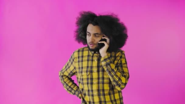 A young man with an African hairstyle on a pink background is talking on the phone. On a colored background — Stock Video