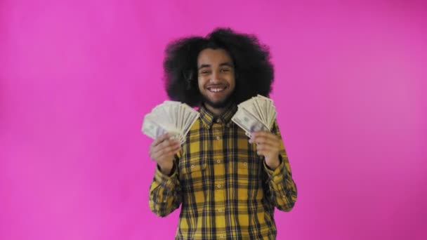 Um homem afro-americano feliz segurando um fã de notas em suas mãos e olhando para a câmera, de pé isolado em um fundo rosa — Vídeo de Stock