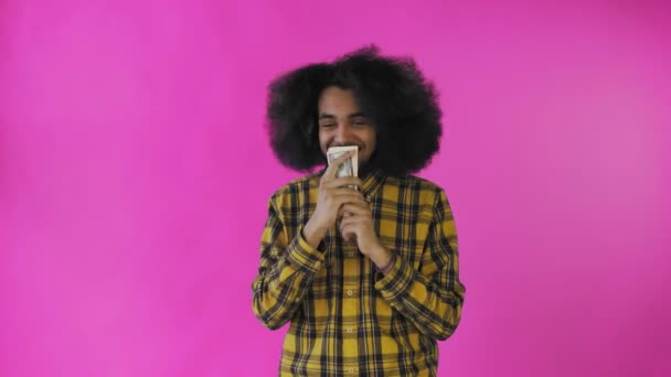 Um homem afro-americano feliz segurando notas em suas mãos, beijando-as e olhando para a câmera, fica isolado em um fundo rosa — Vídeo de Stock