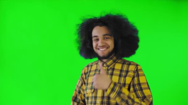 Un hombre afroamericano feliz mirando a la cámara, muestra un parecido con su mano, se encuentra aislado sobre un fondo verde — Vídeos de Stock