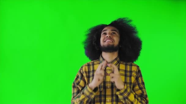 Ein junger Mann mit afrikanischer Frisur auf grünem Hintergrund drückt ihm die Daumen und betet. Emotionen auf farbigem Hintergrund. — Stockvideo