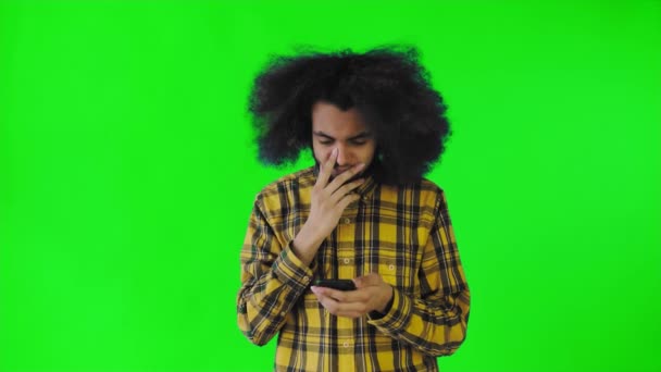 Un giovane con un'acconciatura africana su uno sfondo verde guarda il telefono ed è felicemente sorpreso. Emozioni su sfondo colorato — Video Stock
