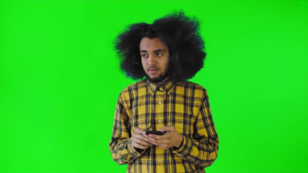 A young man with an African hairstyle on a green background looks at the phone and doubts. Emotions on a colored background — Stock Video