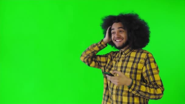 Un giovane con un'acconciatura africana su uno sfondo verde guarda il telefono ed è felicemente sorpreso. Emozioni su sfondo colorato — Video Stock