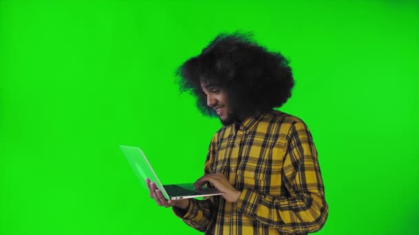 Un joven con un peinado africano sobre un fondo verde está escribiendo en un portátil. Sobre un fondo de color — Vídeos de Stock
