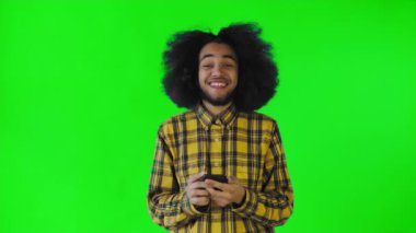 Yeşil arka planda Afrikalı saç stili olan genç bir adam telefona bakar ve mutludur. Renkli bir arkaplanda duygular