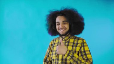 Mutlu bir Afro-Amerikalı adam kameraya bakıyor, eliyle benzer bir şey gösteriyor, mavi arka planda tek başına duruyor.