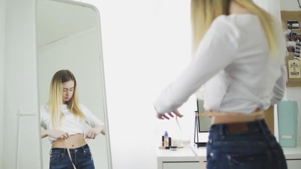 Een jonge vrouw staat voor een spiegel en meet haar taille omtrek met een meetlint — Stockvideo