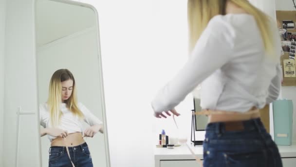 Uma jovem mulher fica na frente de um espelho e mede a circunferência da cintura com uma fita métrica — Vídeo de Stock