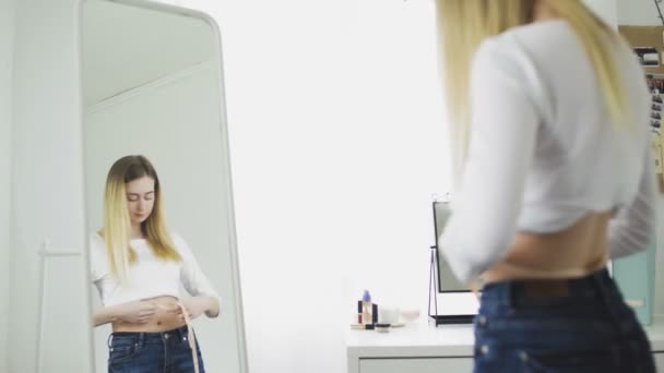 Молода жінка стоїть перед дзеркалом і вимірює окружність талії стрічкою — стокове відео