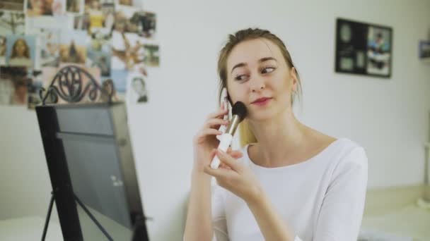 Una giovane donna usa un pennello da trucco per disegnare il rossore sulle guance. La ragazza parla al telefono e si trucca. — Video Stock