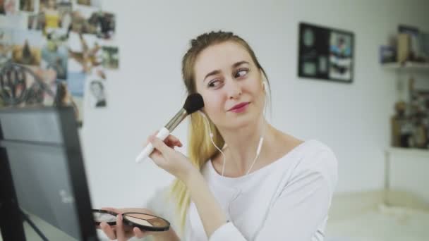 Молодая женщина сидит за столом перед зеркалом, слушает музыку в наушниках и делает макияж — стоковое видео